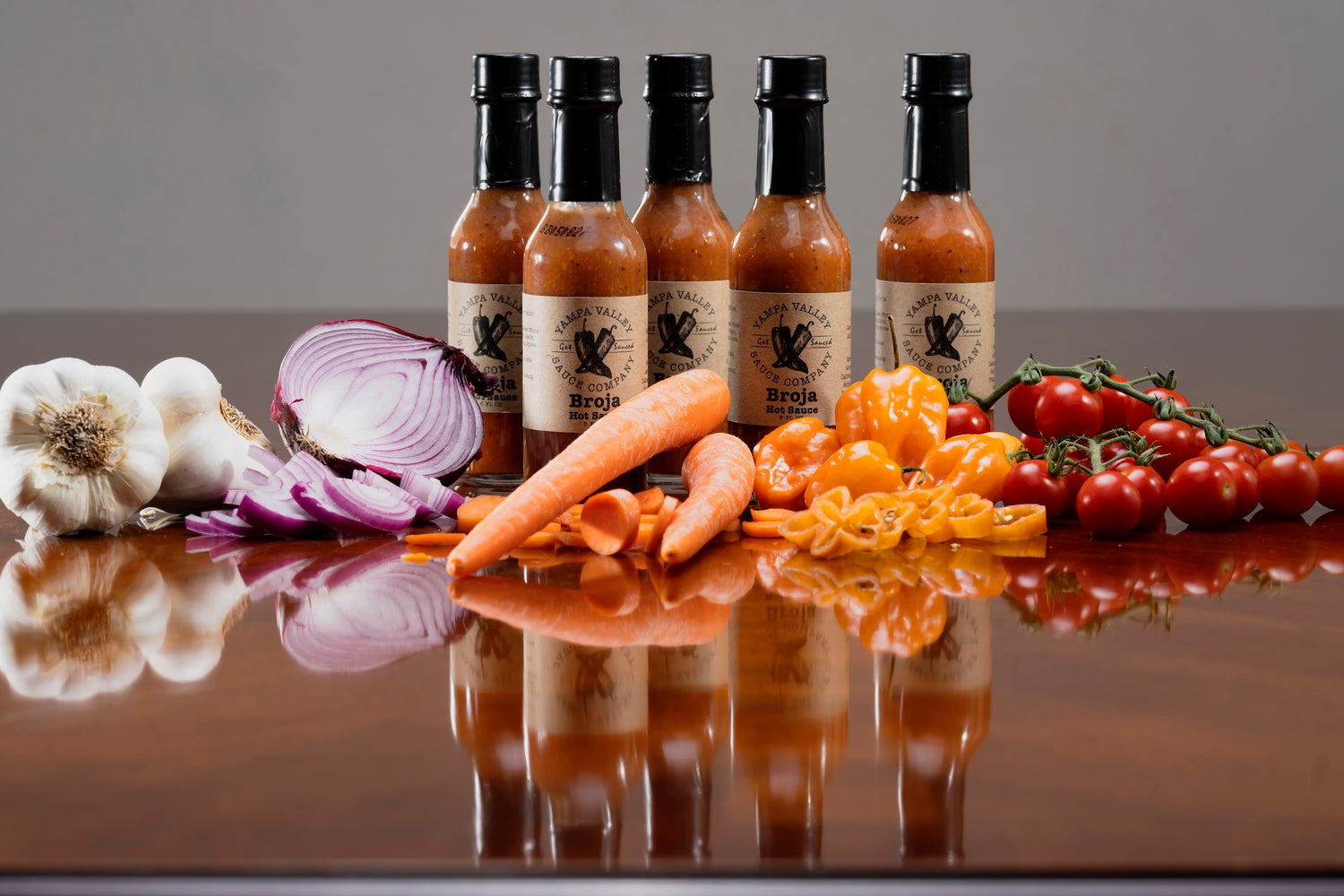garlic, onion, carrot, habañero, and tomato lined up in front of a row of hot sauce bottles