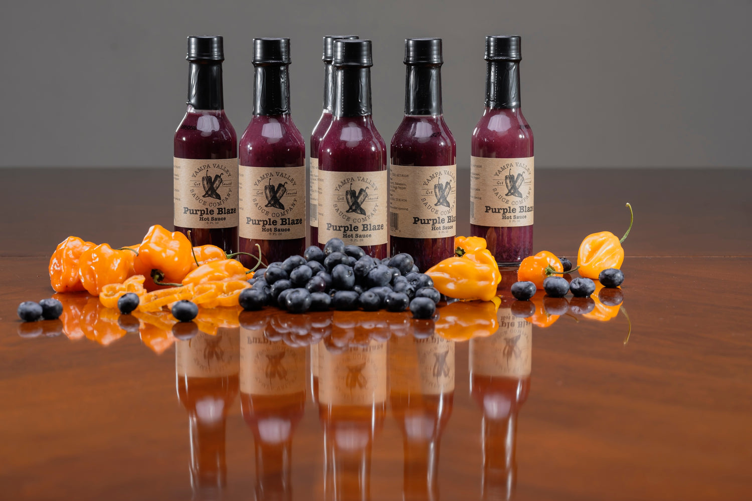 Six bottles of Purple Blaze blueberry hot sauce on a reflective surface with habañero peppers and blueberries.